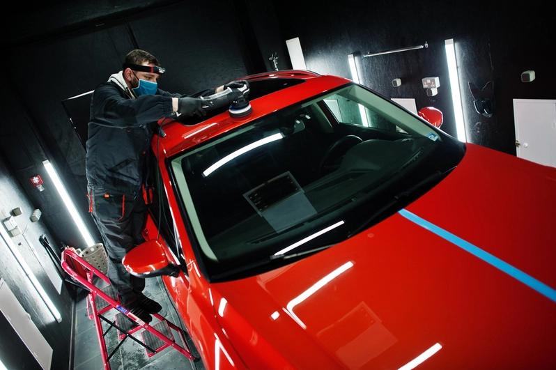 man in face mask with orbital polisher in repair shop polishing roof of orange suv car